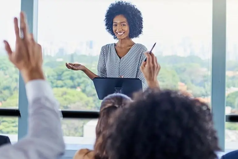 A public speaker smiling while looking at a crowd of business people raising their hands to ask questions